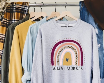 May include: A gray sweatshirt with a rainbow design and the text "SOCIAL WORKER". The rainbow is made up of pink, orange, yellow, and purple stripes with a leopard print pattern in the middle. There is a small heart in the center of the rainbow.