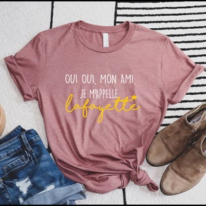 Pu&ograve; includere: Una maglietta rosa con il testo "Oui oui, mon ami! Je m'appelle Lafayette" in lettere bianche e gialle.