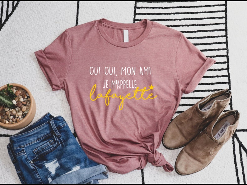 Pu&ograve; includere: Una maglietta rosa con il testo "Oui oui, mon ami! Je m'appelle Lafayette" in lettere bianche e gialle.