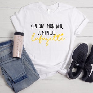 Pu&ograve; includere: Maglietta bianca con testo nero che dice "Oui oui, mon ami, je m'appelle Lafayette" in giallo. La maglietta &egrave; appoggiata su una superficie bianca con un paio di scarpe da ginnastica nere e una tazza da viaggio rosa e bianca.