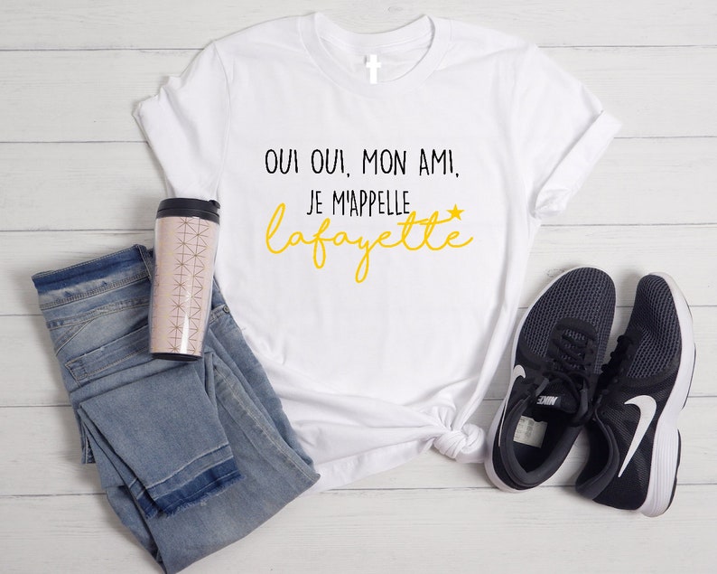 Pu&ograve; includere: Maglietta bianca con testo nero che dice "Oui oui, mon ami, je m'appelle Lafayette" in giallo. La maglietta &egrave; appoggiata su una superficie bianca con un paio di scarpe da ginnastica nere e una tazza da viaggio rosa e bianca.