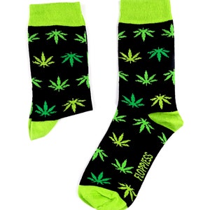 Peut inclure: Une paire de chaussettes noires avec un motif de feuilles de marijuana vertes. Les chaussettes ont un revers vert et le mot "Floppiess" est imprimé sur le côté d'une chaussette.