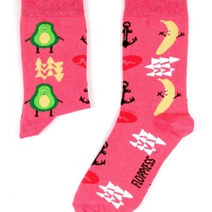 Peut inclure: Une paire de chaussettes roses avec un motif amusant et coloré. Une chaussette présente des avocats, des ancres et des lèvres, tandis que l'autre chaussette présente des bananes, des ancres et des lèvres. Les chaussettes portent l'inscription "Floppiess".