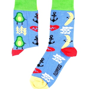 Peut inclure: Une paire de chaussettes bleues avec un motif amusant d'avocats, de bananes, d'ancres et de cœurs rouges. Les chaussettes ont un revers vert et un talon et une pointe jaunes. Les chaussettes sont fabriquées par Floppress.