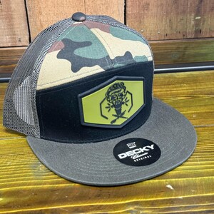 May include: A black and green camouflage trucker hat with a black brim and a yellow patch featuring a cartoon skull with a beard and crossed scissors. The hat has a mesh back and the text "DECKY Classic Original" is printed on the brim.