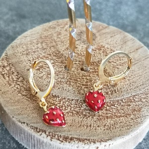 Puede incluir: Un par de pendientes de aro de oro con un dije de fresa roja colgando de cada aro. Los pendientes se muestran en un soporte de madera.