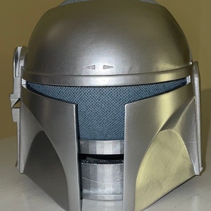 Peut inclure: Un casque argenté avec une grille en maille et une surface texturée. Le casque a un design en relief sur les côtés et un sommet arrondi.