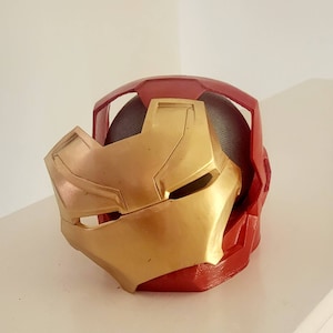 May include: A red and gold 3D printed Iron Man helmet. The helmet is open at the top and the front faceplate is down.