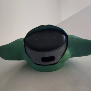 May include: A green and black speaker with a unique design that resembles a character from the Star Wars franchise. The speaker has a round base and two ear-like protrusions.