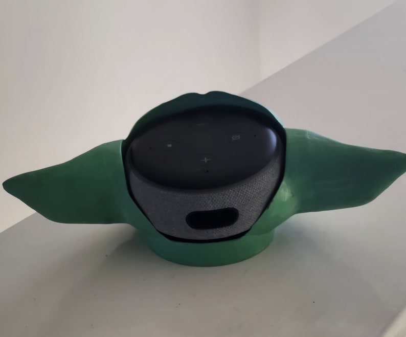 May include: A green and black speaker with a unique design that resembles a character from the Star Wars franchise. The speaker has a round base and two ear-like protrusions.