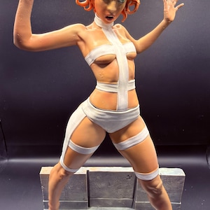 May include: A detailed figurine of a person with orange hair and green eyes, posed with arms raised. The figure wears a white outfit with cross-shaped straps. The sculpture stands on a grey platform with a textured surface, against a dark background.