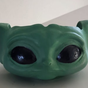 May include: Green ceramic planter shaped like a character from the Star Wars franchise, with large black eyes and two pointed ears.