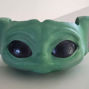 May include: Green ceramic planter shaped like the character Baby Yoda from the Star Wars series. The planter has large eyes and a wide mouth.