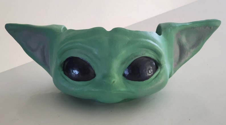 May include: Green ceramic planter shaped like the character Baby Yoda from the Star Wars series. The planter has large eyes and a wide mouth.
