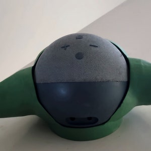May include: A green and grey speaker with a round shape and two small ears. The speaker has a fabric covering and a few buttons on the top.