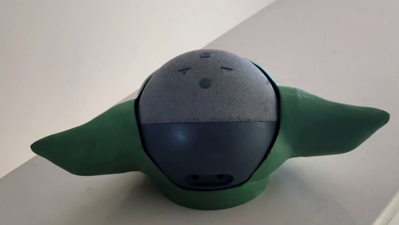 May include: A green and grey speaker with a round shape and two small ears. The speaker has a fabric covering and a few buttons on the top.