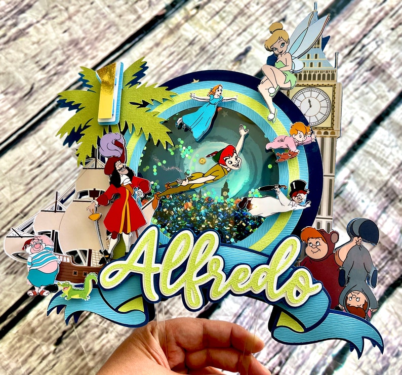 May include: A colorful Peter Pan themed cake topper with a blue and green glitter background. The topper features characters like Peter Pan, Captain Hook, Tinkerbell, and Wendy. The name "Alfredo" is written in green and yellow letters on the topper.