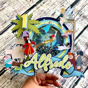 May include: A Peter Pan themed cake topper with a blue and green color scheme. The topper features a ship, a palm tree, a clock tower, and various characters from the movie, including Peter Pan, Captain Hook, Tinkerbell, and the Lost Boys. The name "Alfredo" is written in green and yellow letters on the topper.