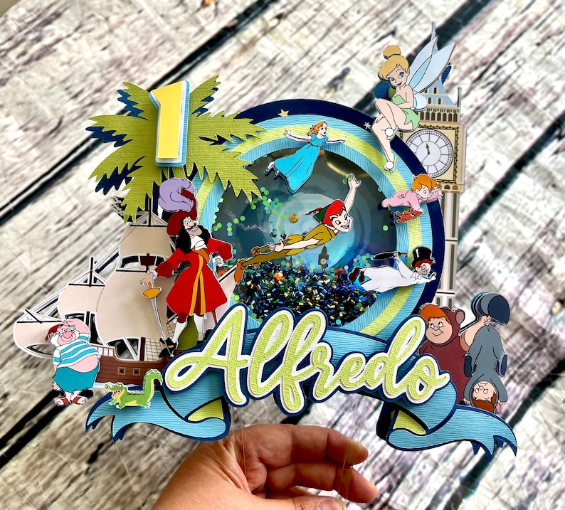 May include: A Peter Pan themed cake topper with a blue and green color scheme. The topper features a ship, a palm tree, a clock tower, and various characters from the movie, including Peter Pan, Captain Hook, Tinkerbell, and the Lost Boys. The name "Alfredo" is written in green and yellow letters on the topper.