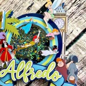 May include: A blue and green cake topper with a glitter background featuring Peter Pan, Tinkerbell, Captain Hook, and other characters from the movie Peter Pan. The topper also includes the name "Alfredo" in green letters.