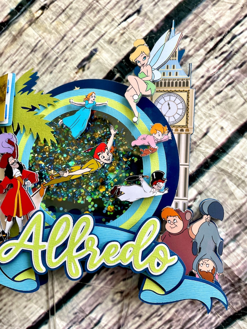 May include: A blue and green cake topper with a glitter background featuring Peter Pan, Tinkerbell, Captain Hook, and other characters from the movie Peter Pan. The topper also includes the name "Alfredo" in green letters.