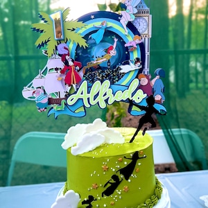 May include: A two-tiered green and white cake with a Peter Pan themed topper. The topper features characters like Peter Pan, Captain Hook, Tinkerbell, and the Lost Boys. The cake is decorated with white clouds and gold stars. The name "Alfredo" is written on the topper.