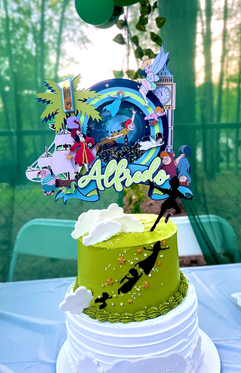 May include: A two-tiered green and white cake with a Peter Pan themed topper. The topper features characters like Peter Pan, Captain Hook, Tinkerbell, and the Lost Boys. The cake is decorated with white clouds and gold stars. The name "Alfredo" is written on the topper.