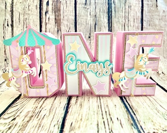 May include: Pink, white, and gold "ONE" letters with a carousel theme. Each letter has a carousel horse, stars, and a pink and teal striped design. The word "Einav's" is written in gold glitter on the "N" letter.