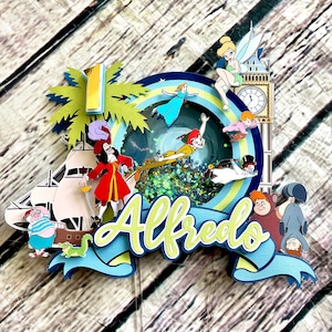 May include: A colorful cake topper featuring characters from Peter Pan and Captain Hook. The topper includes a palm tree, a ship, a clock tower, and a blue and green swirl with glitter. The name "Alfredo" is written in yellow and green letters on a blue ribbon.