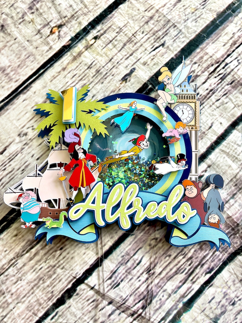 May include: A colorful cake topper featuring characters from Peter Pan and Captain Hook. The topper includes a palm tree, a ship, a clock tower, and a blue and green swirl with glitter. The name "Alfredo" is written in yellow and green letters on a blue ribbon.