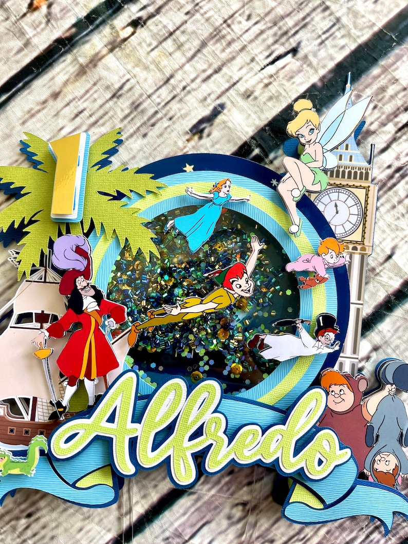 May include: A colorful cake topper featuring characters from Peter Pan, including Peter Pan, Captain Hook, Tinkerbell, and Wendy. The topper also includes a palm tree, a clock tower, and the name "Alfredo".