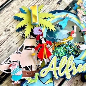 May include: A multi-layered cake topper featuring characters from Peter Pan and Captain Hook. The topper includes a pirate ship, a palm tree, a crocodile, Tinkerbell, Captain Hook, Peter Pan, and a blue and green banner with the name "Alfred".