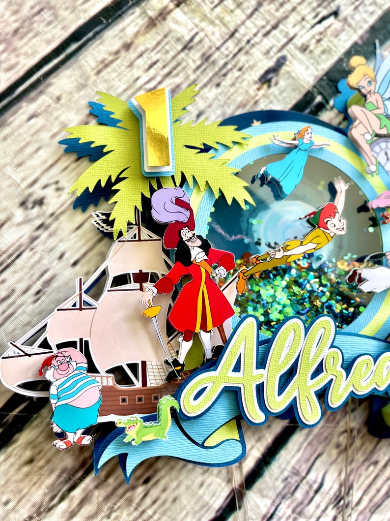May include: A multi-layered cake topper featuring characters from Peter Pan and Captain Hook. The topper includes a pirate ship, a palm tree, a crocodile, Tinkerbell, Captain Hook, Peter Pan, and a blue and green banner with the name "Alfred".