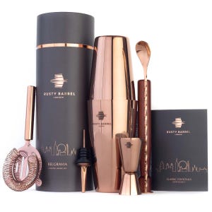 Pode incluir: Um conjunto de mistura de cocktails com ferramentas de bar cor de cobre e uma caixa cilíndrica cinzenta escura. O conjunto inclui um shaker, um coador, um jigger, uma colher e um bico. A caixa está rotulada "Rusty Barrel London".