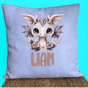 Può includere: Un cuscino blu chiaro con un simpatico drago di cartone animato con fiori rosa e il nome Liam stampato in marrone.