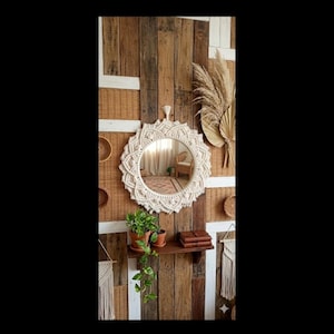 May include: A round mirror with a macrame frame, mounted on a wooden wall with woven panels. A small shelf with plants is below the mirror. The decor has a bohemian style, with natural textures and tones.