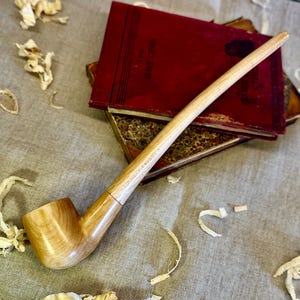 Op de afbeelding: Een lange, houten rookpijp met een gebogen steel en een kom. De pijp is gemaakt van lichtbruin hout en heeft een gladde afwerking. De pijp ligt op een beige stoffen oppervlak met houtkrullen eromheen.