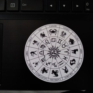 以下が含まれることがあります： 黒色の星座のシンボルと名前が書かれた白い円形のステッカー。シンボルは顔のある太陽の周りに円形に配置されています。星座は、牡羊座、牡牛座、双子座、蟹座、獅子座、乙女座、天秤座、蠍座、射手座、山羊座、水瓶座、魚座です。