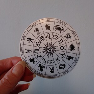 以下が含まれることがあります： 黒色の星座のシンボルと名前が書かれた白い円形のステッカー。シンボルは太陽を中心とした円形に配置されています。星座の名前は円の周りに書かれています。