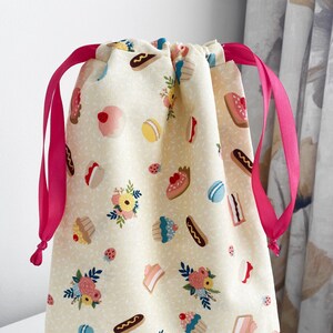 May include: A yellow drawstring bag with a pink ribbon. The bag is decorated with a pattern of cupcakes, pastries, and flowers.