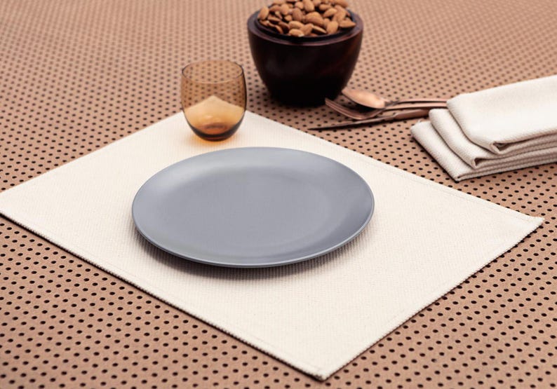 May include: A white placemat with a gray ceramic plate on top. The placemat is on a brown surface with a pattern of small holes. There is a glass of water and a bowl of almonds in the background.