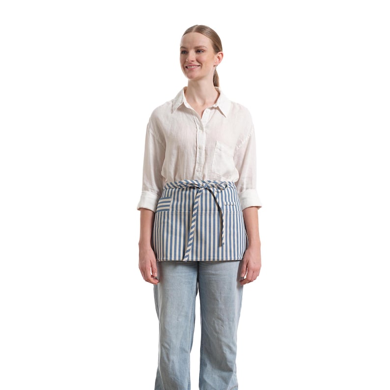 Puede incluir: Una mujer con una camisa de lino blanca y un delantal a rayas azules y blancas. El delantal tiene un bolsillo en la parte delantera y est&aacute; atado a la cintura.