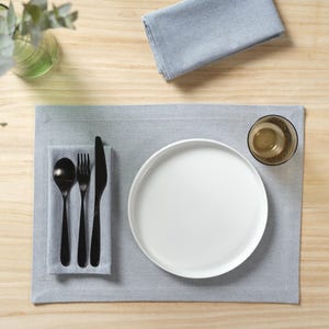 May include: A white dinner plate, black fork, knife, and spoon, and a clear glass on a gray placemat. The placemat is on a light wood surface.