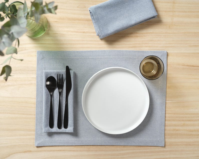 May include: A white dinner plate, black fork, knife, and spoon, and a clear glass on a gray placemat. The placemat is on a light wood surface.