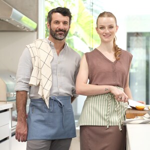 Puede incluir: Un hombre y una mujer con delantales est&aacute;n de pie en una cocina. El hombre lleva un delantal azul claro con una toalla a cuadros blancos y negros sobre su hombro. La mujer lleva un delantal a rayas verde claro y blanco sobre una falda marr&oacute;n. Ella sostiene un cuchillo y una tabla de cortar con naranjas encima.