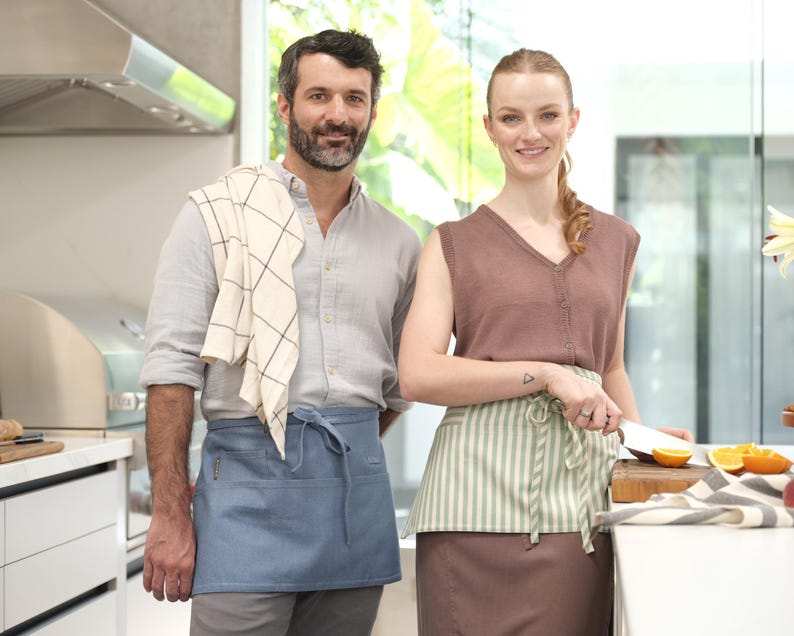 Puede incluir: Un hombre y una mujer con delantales est&aacute;n de pie en una cocina. El hombre lleva un delantal azul claro con una toalla a cuadros blancos y negros sobre su hombro. La mujer lleva un delantal a rayas verde claro y blanco sobre una falda marr&oacute;n. Ella sostiene un cuchillo y una tabla de cortar con naranjas encima.