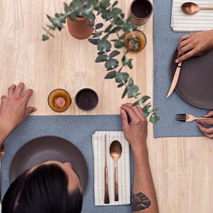 May include: Two people are seated at a wooden table with place settings. Each place setting includes a brown plate, a copper fork, a copper spoon, and a copper knife. The table is set with blue and white striped placemats.