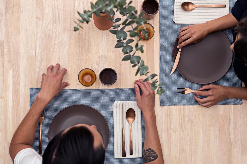 May include: Two people are seated at a wooden table with place settings. Each place setting includes a brown plate, a copper fork, a copper spoon, and a copper knife. The table is set with blue and white striped placemats.