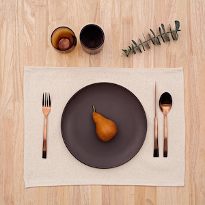 May include: A brown ceramic plate with a pear on a beige placemat. The place setting includes a copper fork and two spoons.