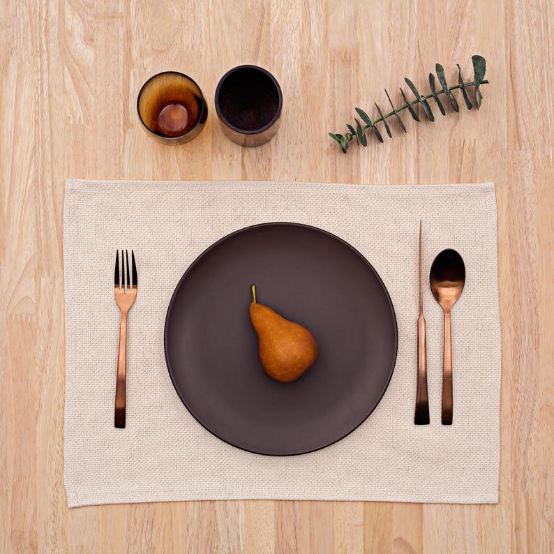 May include: A brown ceramic plate with a pear on a beige placemat. The place setting includes a copper fork and two spoons.